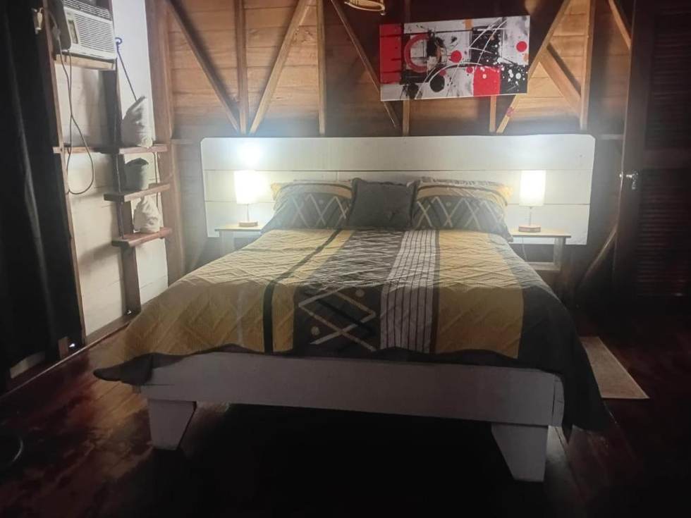 The Double Room: a double bed with a white headboard between two lamps, air conditioning on the wall and a timber-framed ceiling above.