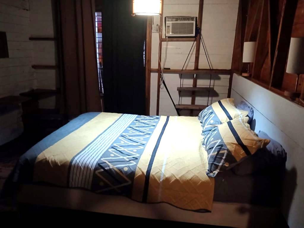 The Double Room by lamplight, the bed made up with a blue and yellow quilt, air conditioning above and shelving along the wall.