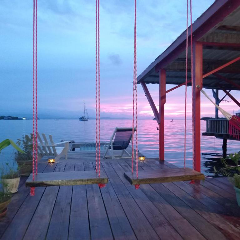 The dock at Villa Bahía del Sol at dusk: two rope swings over a wooden deck, deck chairs facing the water, sailboats moored in Saigon Bay and a pink and violet sky.