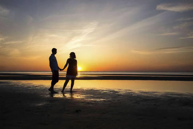 Two people holding hands on wet sand, silhouetted against a low orange sunset over the water.