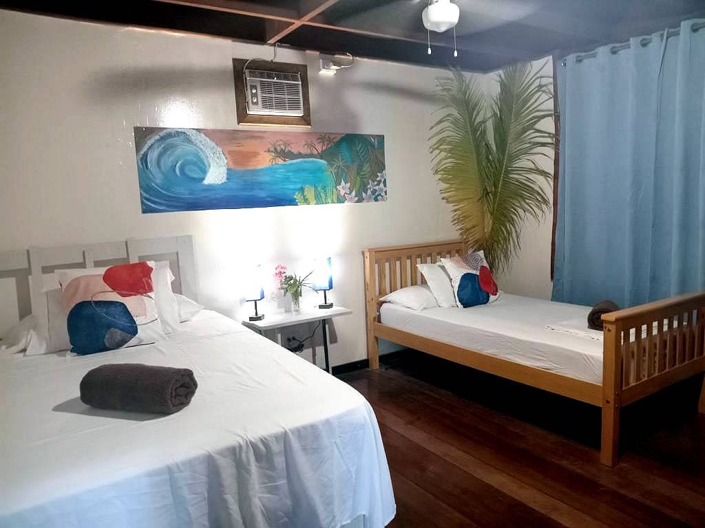 The Twin Room: a large bed and a single bed under a painted wave panel, with air conditioning, a plant in the corner and a curtain across the far wall.